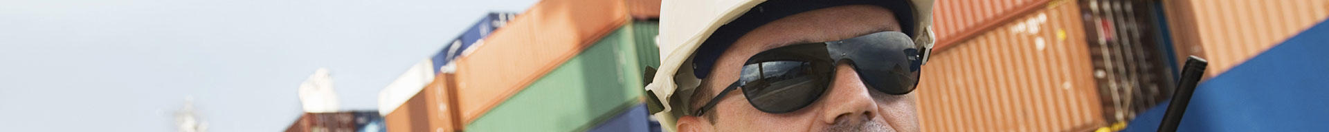 A man wearing a hard hat and sunglasses with shipping containers in the background