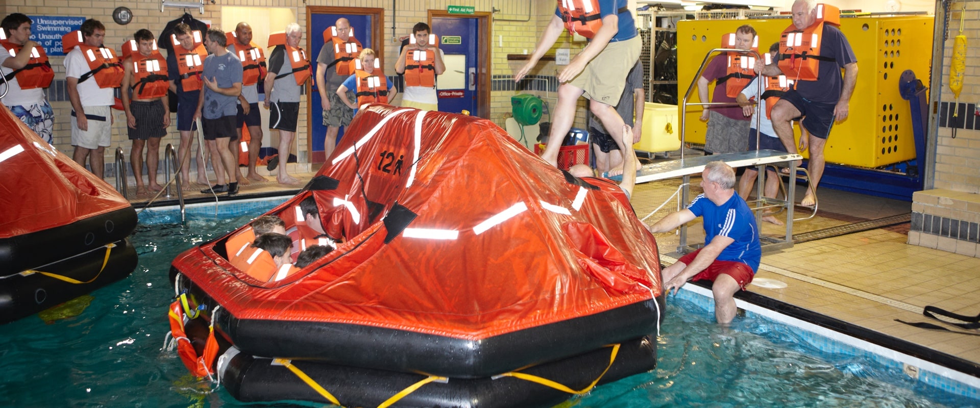 Seafarers in clothes undertaking safety training in swimming pool with life raft